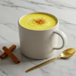 A creamy cup of turmeric milk in a white mug, garnished with cinnamon, with a gold spoon and cinnamon sticks on a marble countertop.