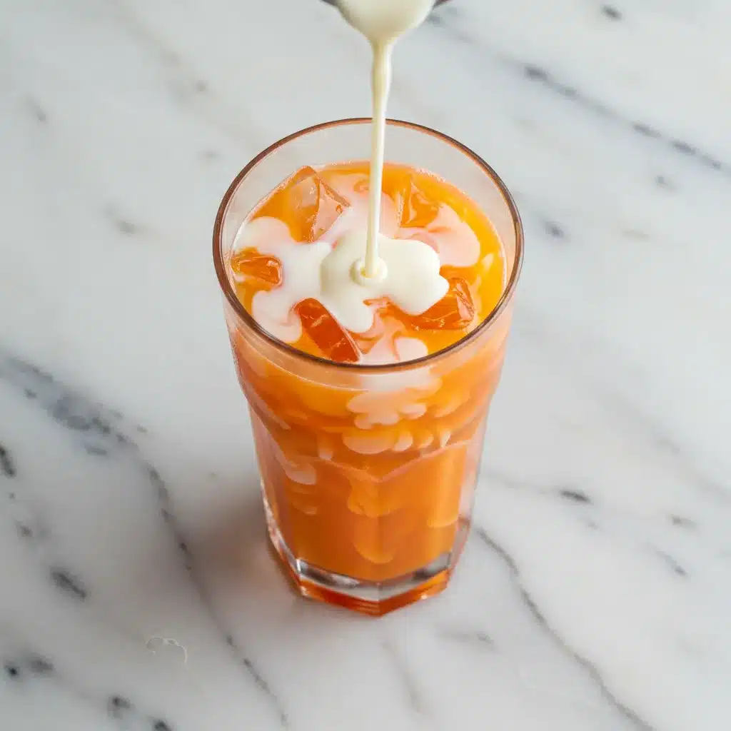 Thai Tea Recipe (Authentic & Easy) 6 An overhead view of thick, sweetened condensed milk being poured into a tall glass of iced Thai tea on a marble countertop.