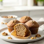 Three moist banana bread muffins on a white plate, with one broken open to show the fluffy texture.