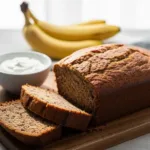 A loaf of banana bread recipe with sour cream, sliced to show the moist interior, with bananas in the background.