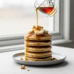 A tall stack of fluffy banana pancakes on a plate, with maple syrup being poured over them and garnished with fresh banana slices.