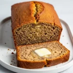A thick slice of the best banana bread recipe resting against the golden-brown loaf on a white platter, showcasing its moist texture.