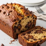 A moist slice of chocolate chip banana bread leaning against the full loaf, showing the gooey chocolate inside.