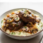 A close up 45-degree angle shot of crockpot salisbury steak served over mashed potatoes in a white bowl.
