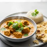A bowl of creamy, golden chicken curry garnished with fresh cilantro, served with basmati rice and naan bread.