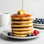 A tall stack of fluffy pancakes with a pat of melting butter and maple syrup being drizzled over the top.