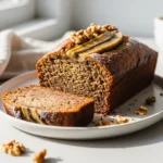 A sliced loaf of moist banana bread on a white platter, with one slice featured prominently to show its texture.
