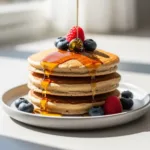 A stack of fluffy vegan pancakes with maple syrup dripping down the side and fresh berries on top.