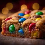A close-up macro shot of a freshly baked M&M Christmas cookie bar, showing the chewy texture and melted chocolate chips alongside colorful candy.