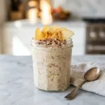 A glass jar of creamy apple cinnamon overnight oats topped with fresh apple slices and pecans.