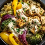 A close-up overhead view of a healthy chicken and veggie bowl with lemon tahini sauce being drizzled over the top. The focus is on the texture of the roasted vegetables and chicken.
