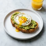 A high protein avocado egg toast on a white plate, topped with a perfectly cooked sunny-side-up egg and garnished with red pepper flakes.