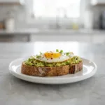 A close up of sourdough avocado toast with a perfectly fried egg on a white plate.