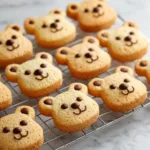 A high-angle view of freshly baked teddy bear cookies cooling on a wire rack, with some chocolate chips visible.
