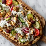 An overhead close-up shot of a vibrant Greek chicken flatbread. The image showcases the colorful toppings like red onions, tomatoes, and herbs.