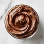 An overhead shot of a glass bowl filled with rich, dark chocolate mousse filling, with a spatula resting on the side showing the smooth texture.