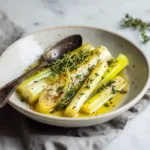 A serving of tender buttered leeks on a white plate, showcasing the simple and elegant final dish. The leeks are glistening and perfectly cooked.