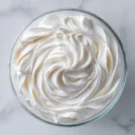 An overhead shot of a glass bowl filled with smooth, whipped Swiss meringue, demonstrating the 'stiff peaks' stage before the butter is added.