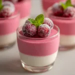 A beautiful clear glass mini dessert cup filled with off-white vanilla mousse and topped with equal thickness of pink cranberry mousse.