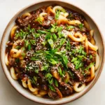 Close-up overhead shot of thick udon noodles tossed with thinly sliced seared beef strips in a sticky sauce.