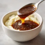 A silver spoon resting inside a bowl, dripping a thick ribbon of smooth medium-brown gravy surrounded by fluffy mashed potatoes.