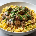 A shallow white ceramic bowl filled with a thick bed of wide egg noodles, topped with tender slow-cooked beef, cremini mushrooms, and creamy brownish-beige gravy.