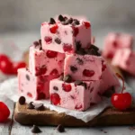 A stack of thick Cherry Garcia Fudge squares on a rustic wooden board. The fudge is pale pink with visible chunks of red maraschino cherries and dark chocolate chips.
