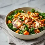 A close-up shot of a vegetarian bowl filled with roasted sweet potato cubes, pan-fried halloumi, and fresh kale, ready to be eaten.
