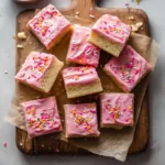 An overhead shot of soft sugar cookie bars cut into neat squares on a white surface, showcasing their vibrant pink frosting and festive sprinkles.