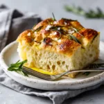 A thick, square slice of sourdough discard focaccia showcasing its airy, open crumb structure and chewy texture. The golden-brown crust is visible, highlighting the successful bake of this no-knead bread.