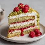 A stunning whole Pistachio Raspberry Cake encrusted with coarse roasted pistachios and crowned with plump red raspberries on a simple white plate.