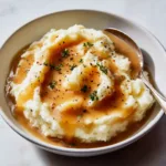Close-up of a white ceramic bowl showing creamy white mashed potatoes topped with glossy brown Copycat KFC Gravy.