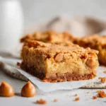Cross section of a sliced Butterscotch Bar on a rustic white ceramic plate, showing fudgy texture and scattered warm baked crumbs.