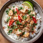 Top-down view of a Coconut Chicken Rice Bowl showing pan-seared chicken coated in creamy white sauce, sprinkled with cilantro and sliced red chilies.