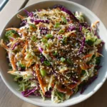 A handheld smartphone photo of an Asian Cabbage Salad in a white bowl featuring green cabbage, purple cabbage, carrots, and raw broccoli florets coated in glossy dressing.