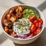High-angle shot of a white ceramic bowl filled with char-grilled chicken cubes and fresh Mediterranean vegetables. (Greek Chicken Marinade)