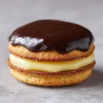 A handheld smartphone photo showing a perfectly stacked Boston Cream Pie Cookie with a soft golden brown base and thick pale yellow cream.