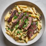 Close-up of Creamy Steak Pasta showing the thick, glossy off-white sauce coating penne pasta and topped with seared steak slices.
