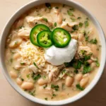 A close-up overhead shot of a white ceramic bowl filled with thick, opaque, pale-cream colored Cream Cheese White Chicken Chili resting on a plain neutral wooden surface in soft natural daylight.