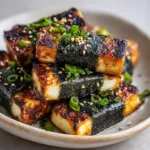 Rustic light-colored ceramic plate piled with pan-fried rectangular tofu blocks snugly wrapped in nori seaweed bands.