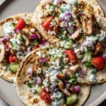 Handheld photo of three Mediterranean Chicken Tacos on a ceramic plate with golden-brown chicken and vibrant vegetables. (Mediterranean Chicken Tacos)