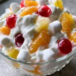Handheld smartphone photo of a glass bowl filled with creamy Ambrosia Fruit Salad with bright orange mandarin segments and red cherries. (Ambrosia Fruit Salad)