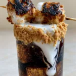 Close up of square ice cubes and dark Coca-Cola with a thick layer of heavy cream cascading down the interior glass walls. (Campfire Dirty Soda)