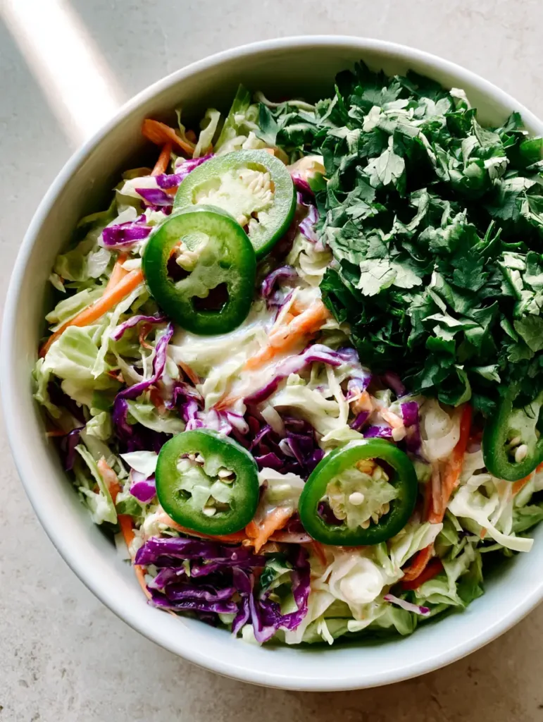 Top-down close-up photo of fresh Jalapeño Slaw with green and purple cabbage and bright green jalapeño rounds in a white ceramic bowl.