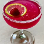 Close up of an elegant coupe glass with a Raspberry Lemon Drop Cocktail featuring a sugar rim and floating lemon wheel.