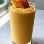 Overhead view of a thick Peach Smoothie With Frozen Peaches showing velvety pale orange texture and peach garnish.