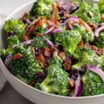Close up of small raw vibrant green broccoli florets mixed with crispy brown bacon and purple onion in a glossy white dressing. (Classic Broccoli Salad)