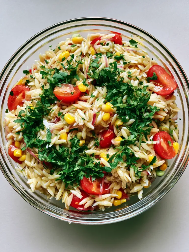 Top-down view of glistening orzo pasta salad with red cherry tomatoes and yellow corn in a clear glass bowl. (Cold BBQ Side Dishes)