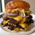 Close up of two thin, jagged-edged smash burger patties with dark brown crusts and melted yellow cheese on a white plate. (Smash Burger Recipe)