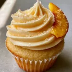 Peach Bellini Cupcakes with Champagne Sponge and Sparkling Frosting Detailed view of the velvety peach-tinted buttercream swirl on a golden-tan champagne sponge cupcake. (Peach Bellini Cupcakes)